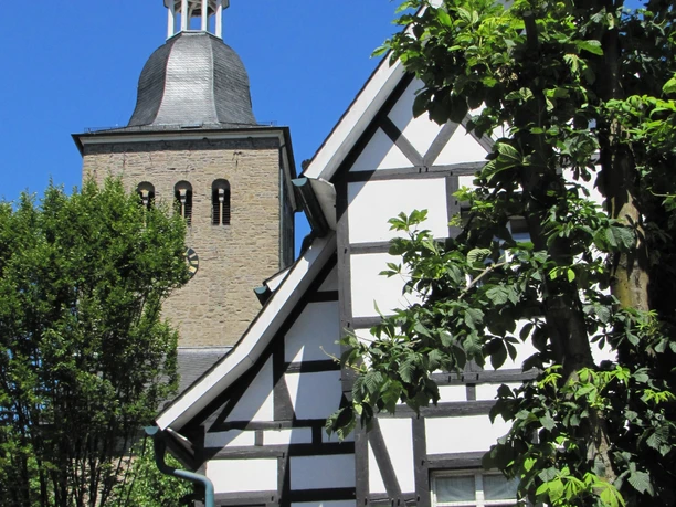 Ortskern Lindlar Fachwerkhaus vor Kirchturm unter blauem Himmel, umgeben von grünen Bäumen.