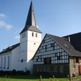 Eine weiße Kirche mit Turm und benachbartem Fachwerkhaus, eingebettet in eine ländliche Landschaft.
