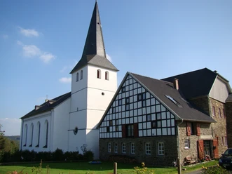 Ev. Kirche und Burg Honrath Eine weiße Kirche mit Turm und benachbartem Fachwerkhaus, eingebettet in eine ländliche Landschaft.