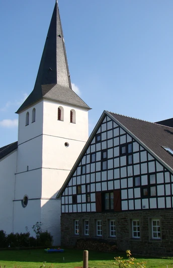 Ev. Kirche und Burg Honrath Eine weiße Kirche mit Turm und benachbartem Fachwerkhaus, eingebettet in eine ländliche Landschaft.