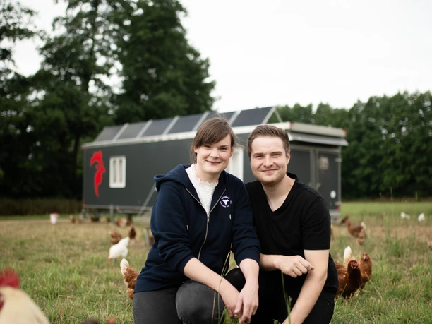 Delbrück_Noltenhof.jpg Two people are sitting on the lawn among the chickens and smiling at the camera