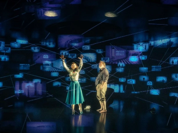 Moneyland - Lavinia Nowak, Benjamin Höppner Zwei Schauspielende in MONEYLAND vor blau leuchtenden Projektionen auf dunkler Bühne