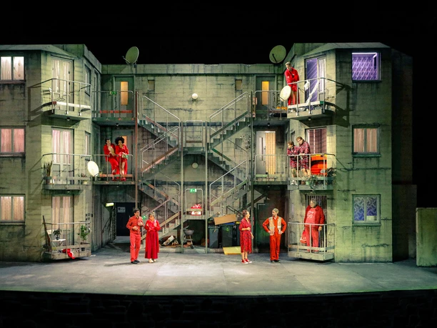 In bester Lage - Ensemble Ensemble in roten Kostümen auf mehrstöckiger Wohnhaus-Bühne im Schauspiel Köln