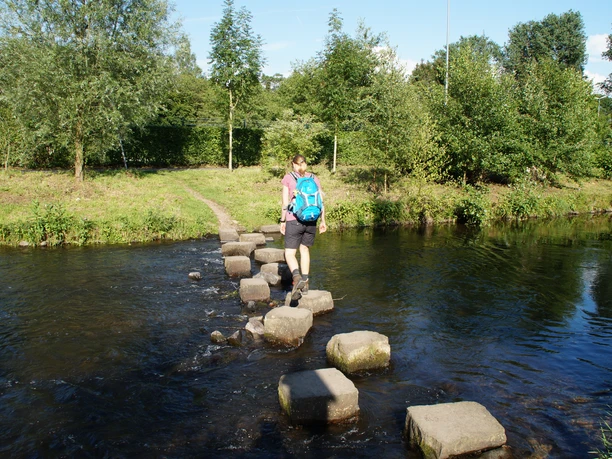 Furt durch die Wupper bei Wipperfürth Eine Frau mit Rucksack überquert auf Trittsteinen einen flachen, klaren Bach inmitten grüner Landschaft.