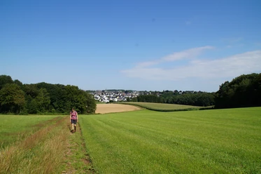 Bergischer Panoramasteig hinter Radevormwald Frau wandert auf einem Feldweg durch grüne Wiesen, mit Blick auf eine Dorfkulisse am Horizont.