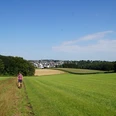 Bergischer Panoramasteig hinter Radevormwald Frau wandert auf einem Feldweg durch grüne Wiesen, mit Blick auf eine Dorfkulisse am Horizont.