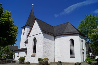 Bonte Kerke in Lieberhausen <p>Weiße Kirche in Gummersbach mit steilem, dunklem Dach vor blauem Himmel und grünen Bäumen.</p>