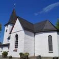 Bonte Kerke in Lieberhausen <p>Weiße Kirche in Gummersbach mit steilem, dunklem Dach vor blauem Himmel und grünen Bäumen.</p>