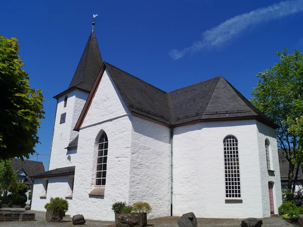 Bonte Kerke in Lieberhausen <p>Weiße Kirche in Gummersbach mit steilem, dunklem Dach vor blauem Himmel und grünen Bäumen.</p>