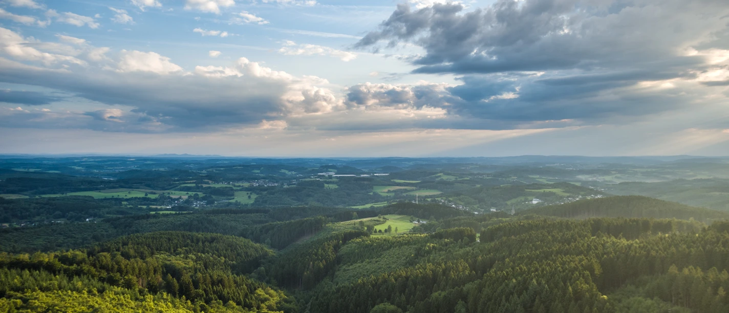 Reichshof Blockhaus Weitläufige, grüne Landschaft mit Wäldern und Hügeln unter einem bewölkten, doch lichtdurchfluteten Himmel.