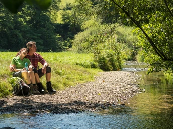 Wandern am Eifgenbachweg Eine Frau und ein Mann sitzen entspannt am Ufer eines ruhigen Bachlaufs in einer grünen Landschaft.