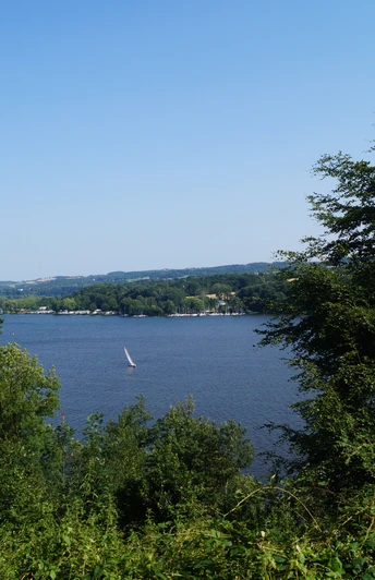 Blick auf den Baldeneysee <p>Blick durch Bäume auf den Baldeneysee in Essen mit Segelboot bei klarem Himmel.</p>