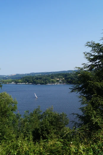 Blick auf den Baldeneysee <p>Blick durch Bäume auf den Baldeneysee in Essen mit Segelboot bei klarem Himmel.</p>