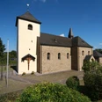 Kirche St. Johann Baptist in Kreuzkapelle Romanische Kirche mit hellem Turm und Kreuz, umgeben von grünem Laub und klarem, blauem Himmel.