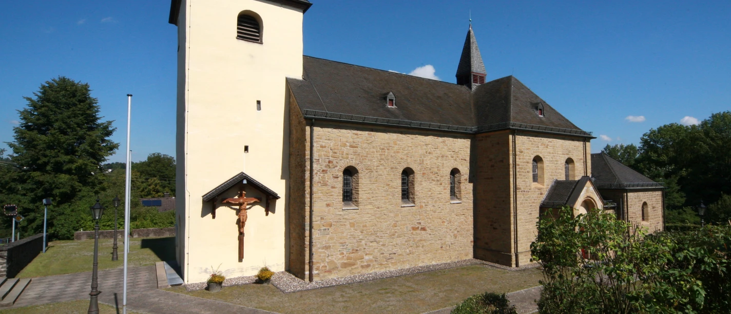 Kirche St. Johann Baptist in Kreuzkapelle Romanische Kirche mit hellem Turm und Kreuz, umgeben von grünem Laub und klarem, blauem Himmel.