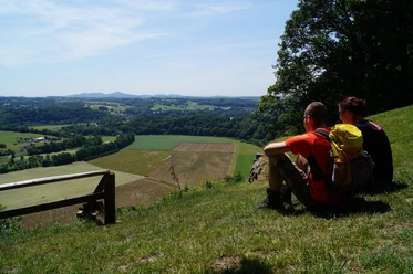 Ausblick vom Stachelberg Paar sitzt auf Wiese, blickt auf weitläufige Landschaft mit Feldern und Hügeln unter klarem Himmel.