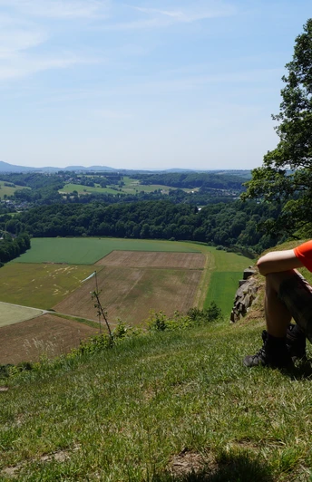 Ausblick vom Stachelberg Paar sitzt auf Wiese, blickt auf weitläufige Landschaft mit Feldern und Hügeln unter klarem Himmel.