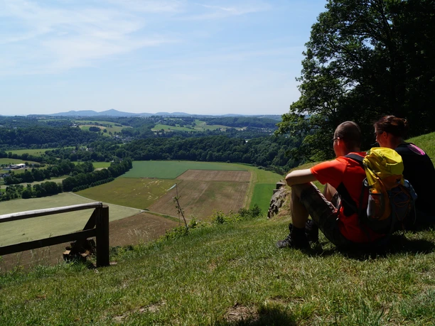 Ausblick vom Stachelberg Paar sitzt auf Wiese, blickt auf weitläufige Landschaft mit Feldern und Hügeln unter klarem Himmel.