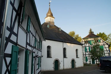 Haan-Gruiten Fachwerkgebäude mit grünen Fensterläden neben einer Kirche mit markantem Turm und Wetterfahne.
