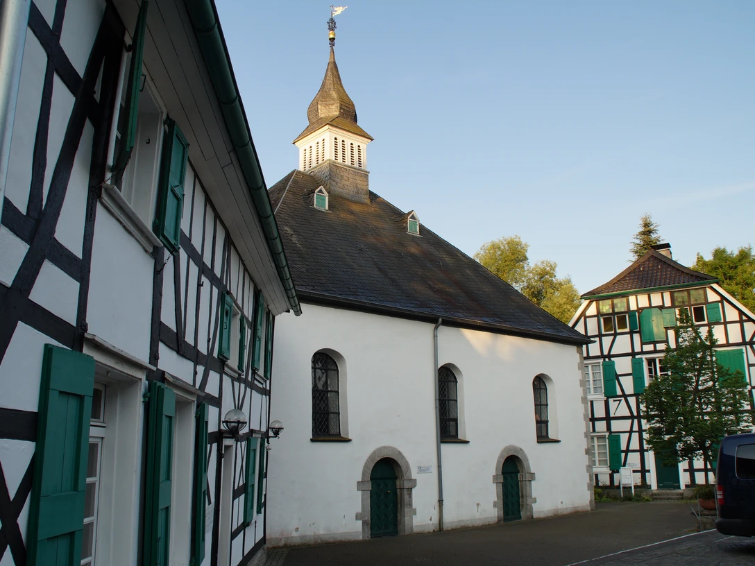 Haan-Gruiten Fachwerkgebäude mit grünen Fensterläden neben einer Kirche mit markantem Turm und Wetterfahne.