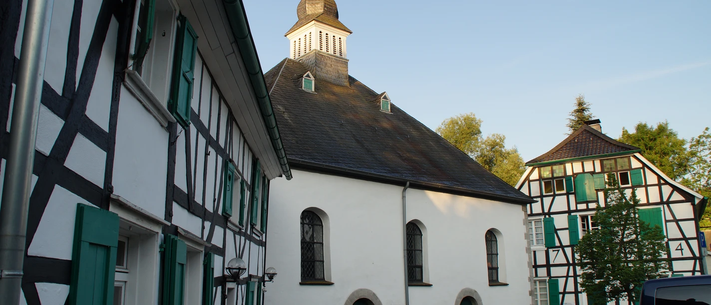 Haan-Gruiten Fachwerkgebäude mit grünen Fensterläden neben einer Kirche mit markantem Turm und Wetterfahne.