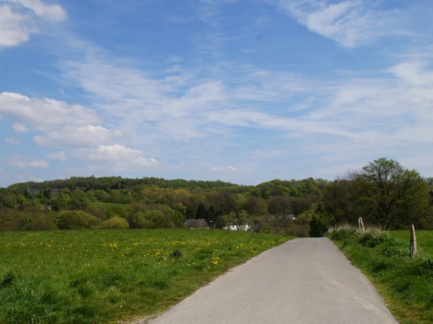 Bergischer Weg Etappe 3 Ländlicher Weg durch grüne Wiesen und Hügel an einem sonnigen Tag mit blauem Himmel und Wolken.