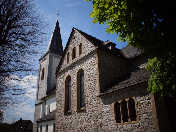 St. Maximin-Kirche in Wülfrath-Düssel Historische Steinkirche mit spitzem Turm und blauer Himmelskulisse, umgeben von Bäumen und Gebäuden.