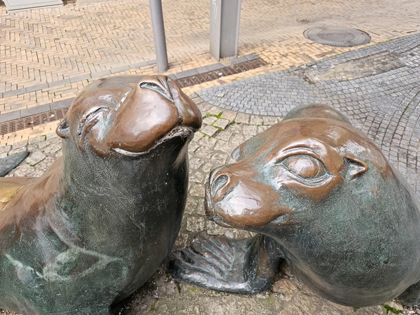 20261015_165733(c) Tanja Mehl_Erlebnis Bremerhaven_WEB.jpg Zwei bronzene Seehundskulpturen an einem Brunnen auf gepflastertem Platz.