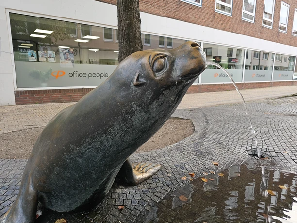 20261015_165519(c) Tanja Mehl_Erlebnis Bremerhaven_WEB.jpg Bronzene Seehundskulptur als Brunnen auf einem gepflasterten Platz in Bremerhaven.