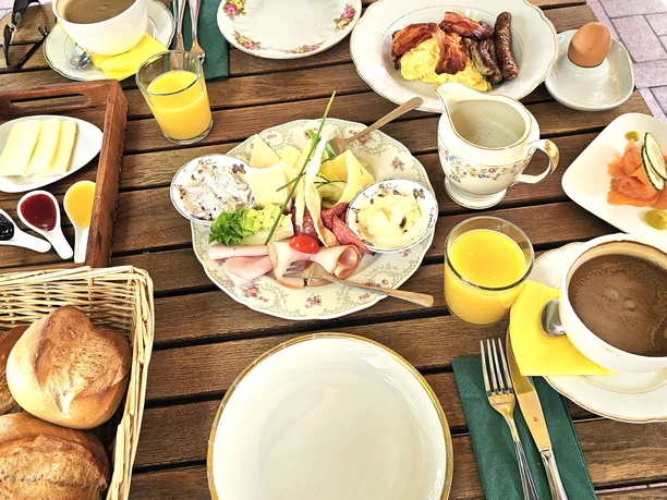 Bistro Lugenstein Breakfast Terrace A breakfast spread on a wooden table featuring rolls, coffee, orange juice, cold cuts, and scrambled eggs.