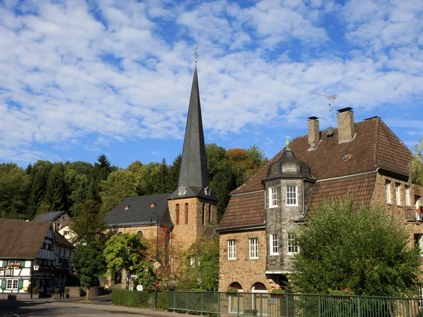 Ortsansicht Eine idyllische Dorfkirche und Fachwerkhäuser unter blauem Himmel mit weißen Wolken.