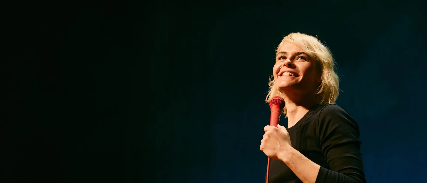 Hazel Brugger Live Hazel Brugger on stage with a red microphone against a dark background