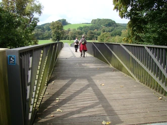 Aggerbrücke am Etappenstart (Gut Eichthal) Eine Holzbrücke führt durch grüne Landschaft; Menschen spazieren, Bäume umgeben den Weg.
