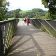 Aggerbrücke am Etappenstart (Gut Eichthal) Eine Holzbrücke führt durch grüne Landschaft; Menschen spazieren, Bäume umgeben den Weg.
