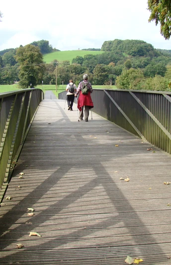 Aggerbrücke am Etappenstart (Gut Eichthal) Eine Holzbrücke führt durch grüne Landschaft; Menschen spazieren, Bäume umgeben den Weg.