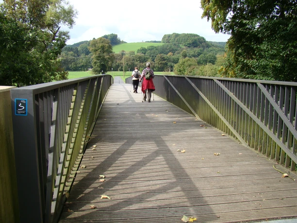 Aggerbrücke am Etappenstart (Gut Eichthal) Eine Holzbrücke führt durch grüne Landschaft; Menschen spazieren, Bäume umgeben den Weg.