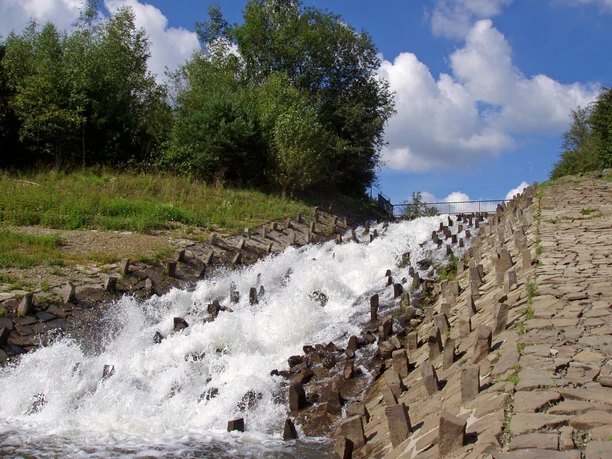 Eindrücke am Wegesrand Wasser fließt über eine steinbefestigte Kaskade, umrahmt von grünem Bewuchs unter blauem Himmel.