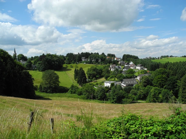 Eindrücke am Wegesrand Panoramablick auf ein grünes Dorf inmitten ländlicher Hügellandschaft unter blauem Himmel.