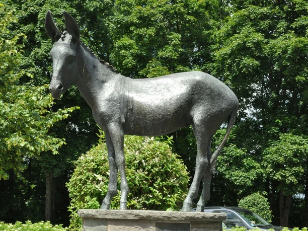 Esel am Kreisel in Kürten Bechen Bronzestatue eines Esels auf einem Sockel, umgeben von üppigem Grün und Bäumen im Hintergrund.