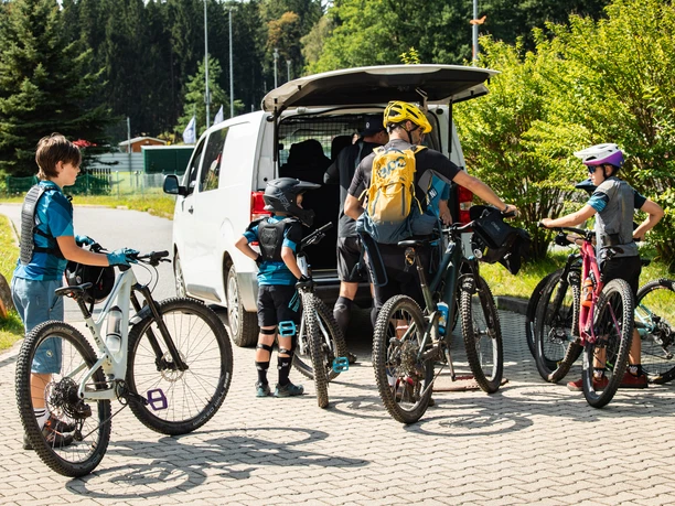 BorderlessTrailsShuttle.jpg Vier Mountainbiker stehen mit ihren Fahrrädern am geöffneten Heck eines weißen Transporters und bereiten sich auf eine gemeinsame Tour vor.