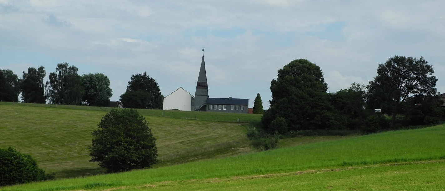 Eindrücke am Wegesrand Kirche mit spitzem Turm in grüner Hügellandschaft unter wolkigem Himmel, umgeben von Bäumen.