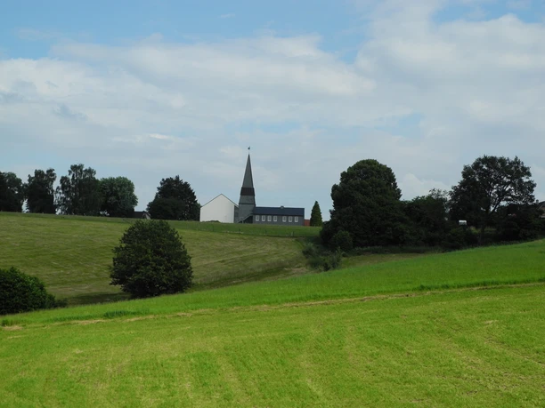 Eindrücke am Wegesrand Kirche mit spitzem Turm in grüner Hügellandschaft unter wolkigem Himmel, umgeben von Bäumen.