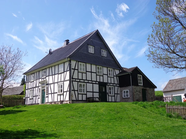 Schultheißmühle Ein traditionelles Fachwerkhaus mit schwarzem Holz und weißen Wänden auf grünem Hügel bei blauem Himmel.