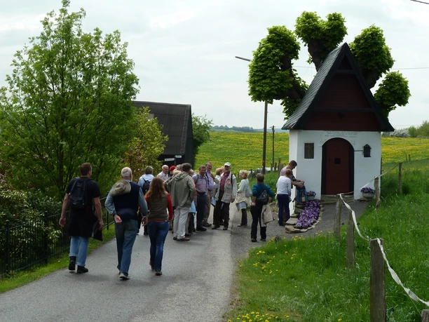 St. Andreas Kapelle in Biesenbach Gruppe von Menschen besichtigt eine kleine Kapelle neben blühenden Feldern und einem grünen Baum.