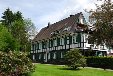 Berger Mühle Historisches Fachwerkhaus mit weißen Wänden, grünen Fensterläden, umgeben von Garten und Bäumen.