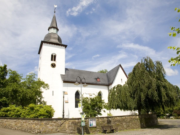Bonte Kerke Marienberghausen Weiße Kirche mit spitzem Turm und Uhr, umgeben von üppigen Bäumen bei strahlend blauem Himmel.