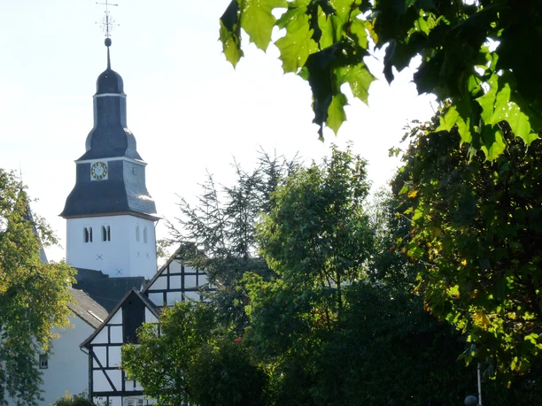 1.000-jährige Schlosskirche in Nümbrecht Eine weiße Fachwerkkirche mit dunklem Turm und Uhr, umgeben von grünen Blättern und Bäumen.