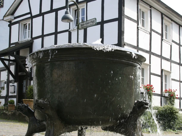 Hexenbrunnen Ein großer, antiker Bronze-Kesselbrunnen in einem gepflasterten Hof vor einem Fachwerkhaus.