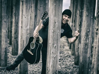 Werner Weimar Schwarz-weiß Aufnahme eines Musiker mit einer Mütze und einer Gitarre in der Hand der hinter einem Holzpfosten hervorschaut.