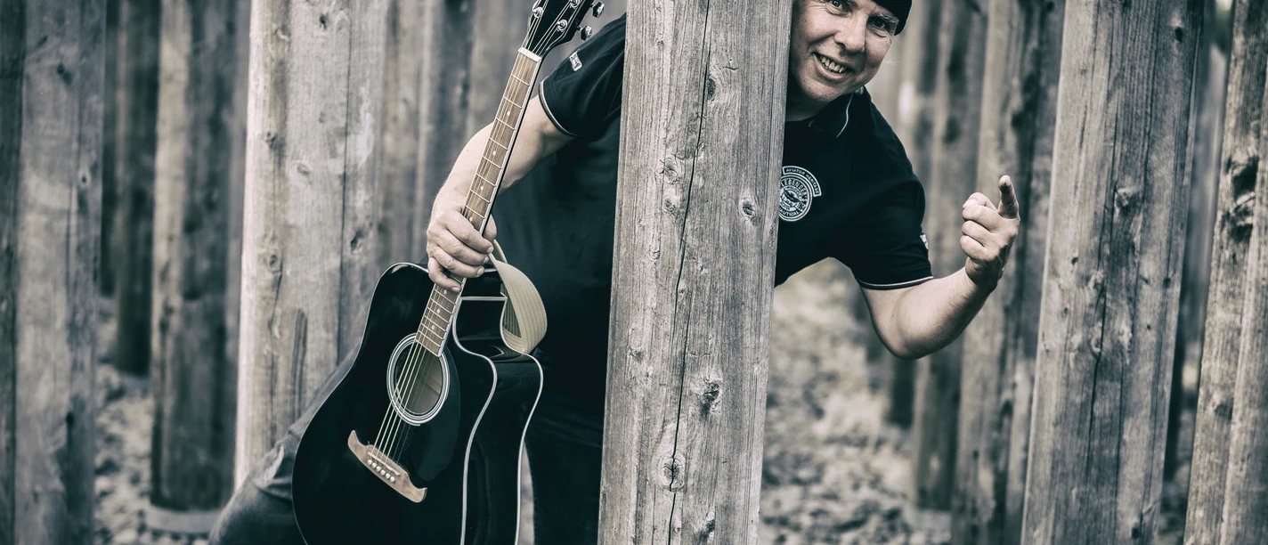 Werner Weimar Schwarz-weiß Aufnahme eines Musiker mit einer Mütze und einer Gitarre in der Hand der hinter einem Holzpfosten hervorschaut.